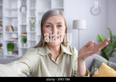 Mature woman communicating virtually, making a video call from her home, smiling and gesturing while looking directly at the camera, staying connected online Stock Photo
