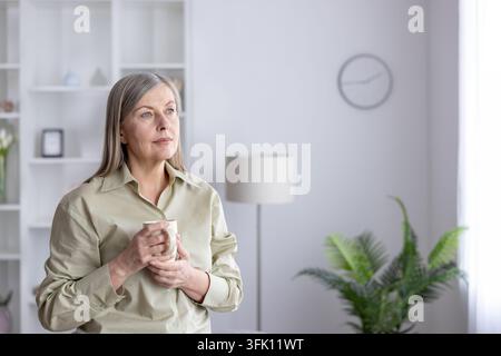 Mature woman with grey hair holding a warm mug, standing in a bright home interior, thoughtfully looking away, reflecting on life's moments with a pensive expression Stock Photo