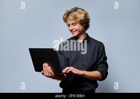 Portrait of smiling young man or student holding laptop computer typing on keyboard while looking at screen on grey background. Remote study e-learnin Stock Photo