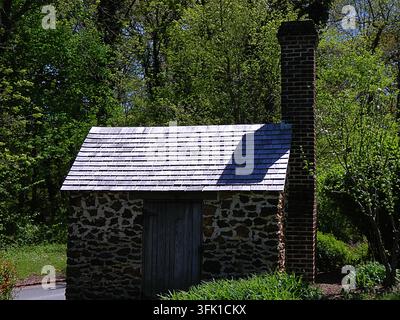 Historic fieldstone outbuilding featuring a weathered wooden shingle roof and a tall brick chimney, set against a dense green forest backdrop. Stock Photo