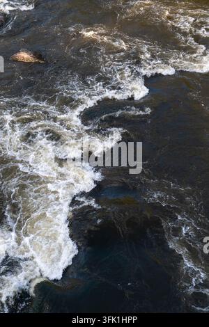 Whitewater rapids sweep diagonally across dark textured rocks, creating frothy white foam beside brown swirling currents in a close overhead view of a Stock Photo