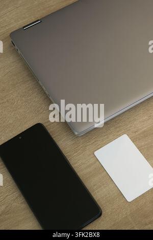 Top view of a closed silver laptop, black smartphone with blank screen, and a plain white digital access card arranged on a wooden desk surface, repre Stock Photo