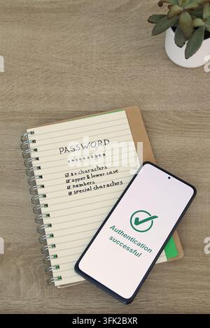 Smartphone showing "Authentication successful" message resting diagonally on a notebook with password strength checklist, on a wooden desk beside a sm Stock Photo
