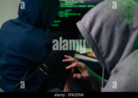 Two anonymous hackers in hoodies working together on computer. Close-up on hand typing on keyboard with green code on screen in background. Concept of Stock Photo
