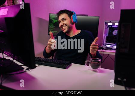 Portrait of smiling young man wearing headphones and giving two thumbs up gesture sitting at desk with multiple computer screens displaying code in Stock Photo