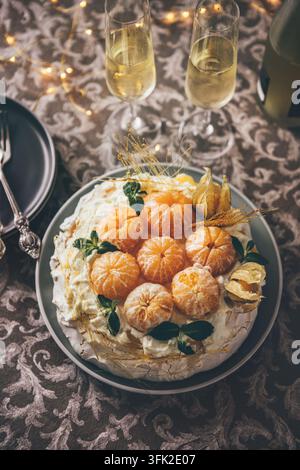 Festive Pavlova meringue cake decorated with fresh mandarins, physalis, mint and caramel served with champagne glasses on elegant table setting during Stock Photo