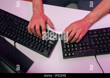 Top view of person hands illuminated by ambient light, actively using two black computer keyboards on bright desk, engaging in work, gaming, coding Stock Photo