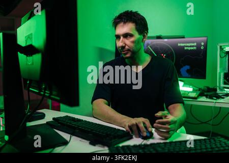 Focused man at desk with multiple screens, engaged in hacking or programming. Dark room with intense green light creates mysterious mood. Concept for Stock Photo