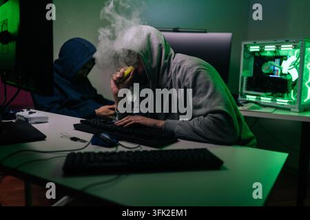 Hooded person vaping and typing on keyboard, engaged in cyber activity alongside another individual, surrounded by glowing computer components in dark Stock Photo