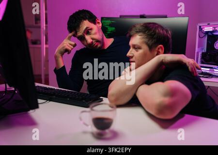Two focused male developers work together at night. One man puzzled by code on screen coworker watches. Moody purple lighting creates tech atmosphere Stock Photo