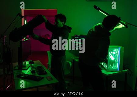 Two technicians prepare for filming or streaming in dark room. Men adjust professional lights near computers with code on screens. Green neon glow Stock Photo