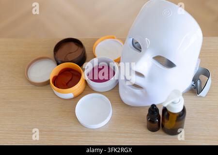 White LED facial mask is resting on light wood tabletop with open cream jars, amber bottles Stock Photo