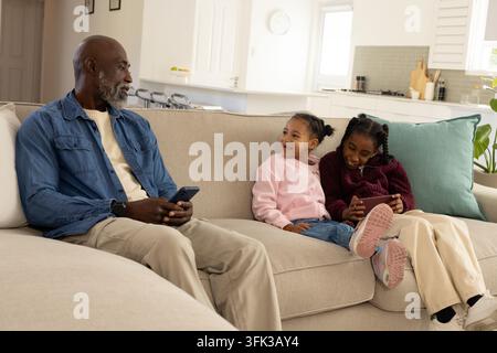Mature African American man with female children sitting on sofa at home holding smartphone, tablet Stock Photo