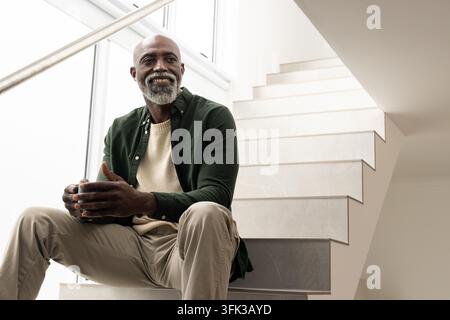 Mature African American man sitting on stairs by window, holding mug, wearing shirt and sweater Stock Photo