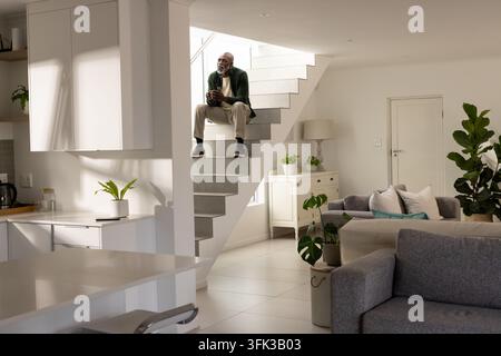 Senior African American man sitting on floating stairs holding mug, looking toward white cabinets Stock Photo