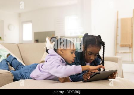 Two African American school-age girls lying on beige sofa pointing and tapping tablet together Stock Photo