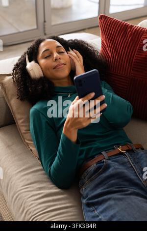 African American woman reclining on sofa at home, wearing teal top, holding phone, touching headset Stock Photo