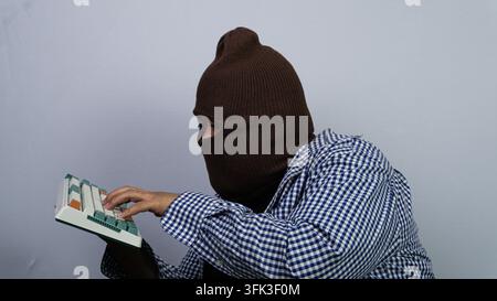 An adult Asian male wearing a brown balaclava mask and a checkered shirt types suspiciously on a mechanical keyboard. The masked individual operates t Stock Photo