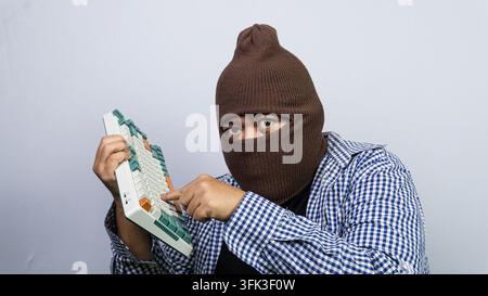 A young Asian male wearing a concealing brown balaclava and a plaid shirt points directly at a mechanical computer keyboard. The disguised subject loo Stock Photo