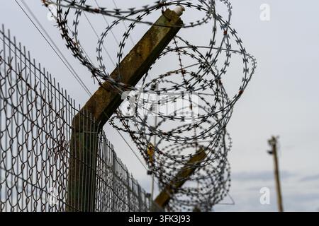 Barbed wire coils tightly around a rusted wooden post. Metal fence stretches behind, gray clouds above. Weathered structure signals security, confinem Stock Photo