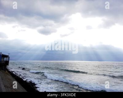 A stunning coastal scene featuring dramatic crepuscular rays breaking through dark clouds over a turbulent sea with rolling waves. A weathered pier st Stock Photo