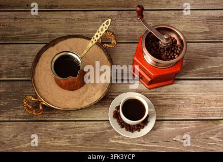 Cezve in a tray with sand, hand coffee grinder with roasted beans and a cup of hot coffee on a wooden table. Top view, flat lay. Stock Photo