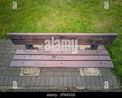 An overhead view of an empty wooden park bench, showing its weathered brown planks and metal frame, situated on paving stones with green grass surroun Stock Photo