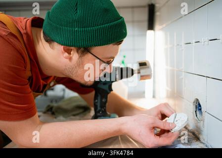 Man using a power drill with a core bit to cut a precise hole in ceramic kitchen wall tile for installing an electrical outlet during a home renovation project Stock Photo