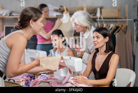 In sewing class for group of beginners, enthusiastic young woman working on machine, stitching fabric pieces of garment and discussing pattern with cl Stock Photo