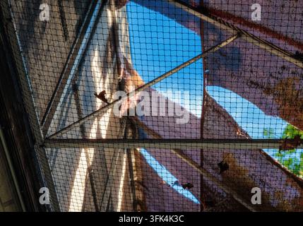 Abstract view of sunlight filtering through a torn fabric canopy metal frame and safety netting creating geometric shadow patterns against blue sky. Stock Photo