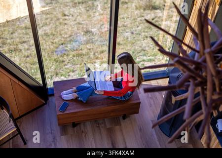 Woman Using Laptop Beside Large Windows in Modern Cabin. Young woman in a red sweater uses a laptop while beside large windows in a modern cabin. Stock Photo