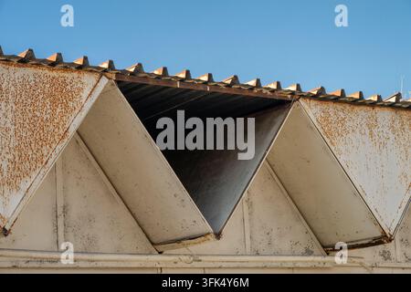 The geometric roofline of an old industrial building displays weathered, rusted triangular folded panels beneath a corrugated rooftop against a clear Stock Photo