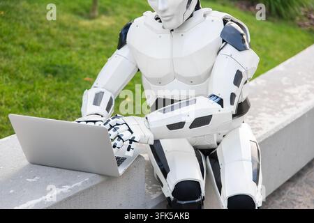 Humanoid robot typing on silver laptop keyboard while sitting on concrete bench outdoors Stock Photo