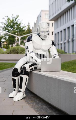 Cybernetic robot working on modern laptop in city park near office building Stock Photo