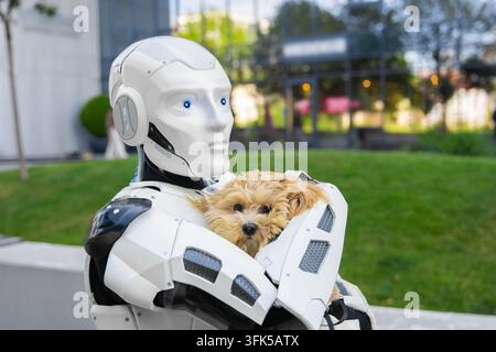 Futuristic white robotic cyborg safely carrying adorable small dog in arms standing in green city park near contemporary corporate architecture Stock Photo