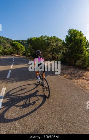 High-angle rear view of a young female cyclist pedaling out of the saddle on her road bike along a paved mountain road, casting a long shadow, on the Stock Photo