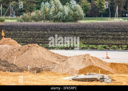Large heaps of excavated sand and soil sit beside concrete slabs and a orange traffic cone at an outdoor road construction site during daytime. A plow Stock Photo