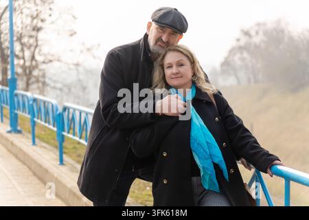 A mature couple embrace beside a blue railing outdoors. A bearded man in a flat cap stands behind a smiling blonde woman wearing a turquoise scarf. Stock Photo