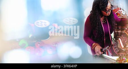 African fashion designer cutting patterned fabric at her workspace, surrounded by sewing materials, clothing and textiles. Stock Photo
