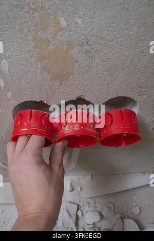 Worker's hand installing multiple red plastic electrical wiring boxes into a recessed hole in a rough concrete wall during a home renovation or constr Stock Photo
