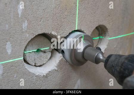 Worker drilling holes in a concrete wall with a powerful rotary hammer and hole saw attachment, using a green laser level for precise positioning duri Stock Photo