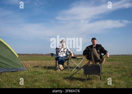Young couple camping in a sunny meadow, sitting by a tent and portable grill, relaxing together under blue skies while enjoying outdoor cooking, bondi Stock Photo