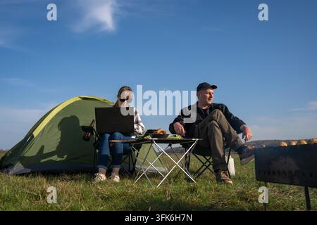Couple camping in a sunny meadow: woman focused on laptop working remotely while man relaxes by the tent, enjoying nature, freedom and a peaceful outd Stock Photo