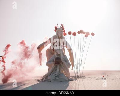 Surreal conceptual performance art featuring two women in white costumes with lace face coverings holding sphere among tall red artificial flowers and Stock Photo