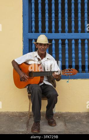 Trinidad, Sancti Spiritus, Cuba - 07 February 2020: View of an elderly man in a straw hat playing an acoustic guitar while sitting on a stool against Stock Photo
