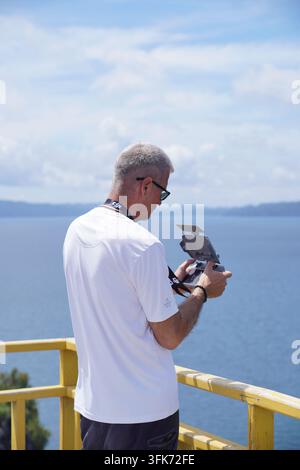 Raja Ampat, Southwest Papua, Indonesia - 06 September 2026: View of a man operating a drone controller on a yellow balcony overlooking the blue waters Stock Photo