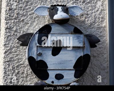 A decorative metal mailbox shaped like a cartoon cow with black and white spots, mounted on a textured stucco wall. Stock Photo