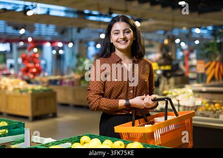Young woman smiling and holding a shopping basket while choosing fresh produce at a supermarket, representing daily consumer lifestyle and retail experience Stock Photo