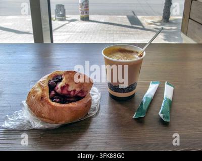 A paper cup of coffee and a sweet berry bun on a wooden table in a cafe in Zaporizhzhia, Ukraine. Two sugar sticks lie nearby Stock Photo