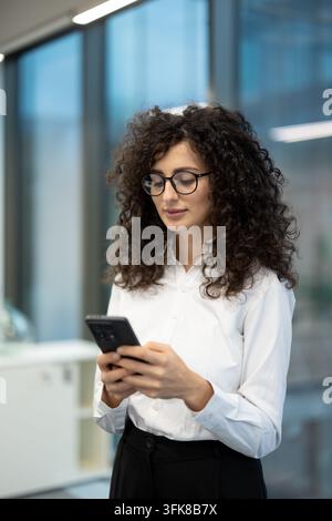 Business communication and digital connectivity concept, young woman using a smartphone for professional tasks in a modern office. Professional employee checking her phone for corporate updates. Stock Photo
